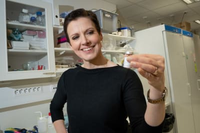 En kvinna står i ett laboratorium med en liten flaska i handen. Hon ser nöjd ut.