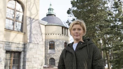 Vaalehiuksinen nainen seisoo kivirakennuksen edustalla ja katsoo suoraan kameraan.