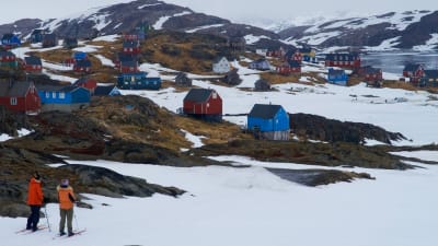 Två personer står på skidor. Blickar ut över en liten by i Grönland. Husen är blåa och röda. 