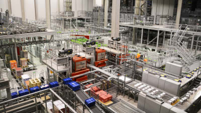 Vy inifrån logistikcentral. En massa rullband och produkter som åker förbi samt några arbetare i orange kläder synns.