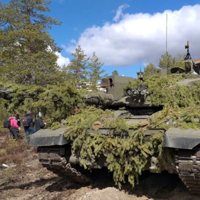 Havuin peitetty panssarivaunu metsäisessä maisemassa. Vasemmalla kuvaajia.