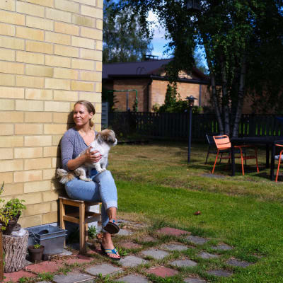 Terhi Vallineva istuu asuntonsa takapihalla puutarhatuolissa. Aurinko paistaa ja Vallineva pitää sylissään Börje-koiraa.