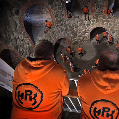 Oranssihuppariset miehet seisovat ylhäällä ja katsovat alapuolellaan olevia pelaajia.