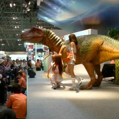 Kauko-ohjattava dinosaurusrobotti ja esiintyjiä teknologiamessuilla.