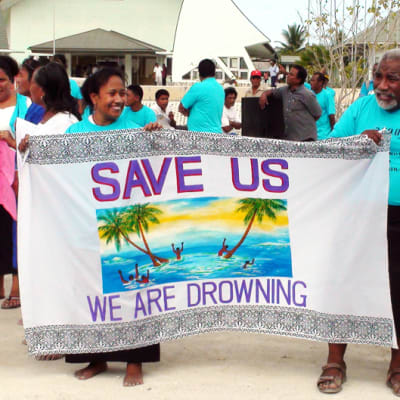 Kiribatin paikallisasukkaita lakanan kanssa. Lakanassa lukee Save us, we are drowning.