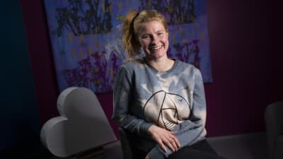 Manusförfattaren sitter på en stol mot en blå-lila bakgrund, håret fäst i en hästsvans och småler rakt mot kameran. Bredvid stolen ligger en sned hjärtsymbol.