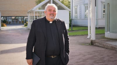 Man i svart kostym och prästkrage står på en innergård.