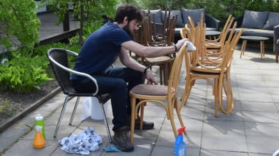 En ung man sitter utomhus på en bakgård och putsar sotiga stolar som har smutsats ner på grund av brand inne i ett restaurangkök.
