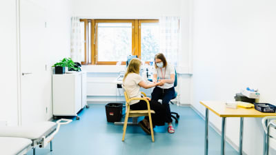 En hälsovårdare sitter med en patient i sitt rum. Hon håller patientens hand, det verkar som om hon undersöker något.