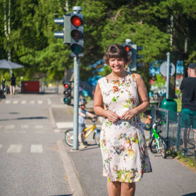 En glad kvinna står på en asfalterad gångbana. I bakgrunden syns ett barn på en cykel och ett rött trafikljus.