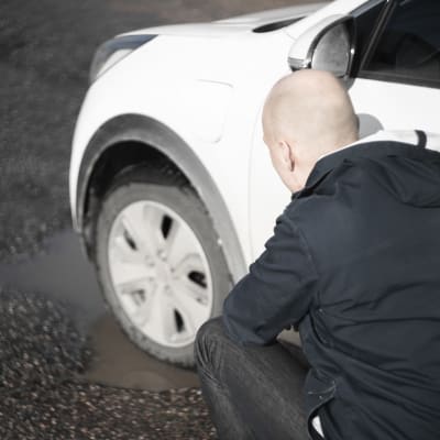 En man står vid en vit bil på en parkeringsplats. Bilens däck är i en vattenfylld grop. Mannen ser oroligt på däcket.