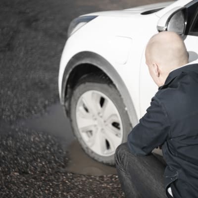 En man står vid en vit bil på en parkeringsplats. Bilens däck är i en vattenfylld grop. Mannen ser oroligt på däcket.