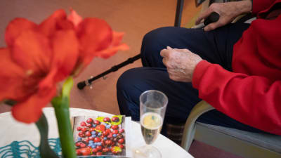 En äldre person sitter med sin promenadkäpp vid ett b ord där det finns ett glas champagne.