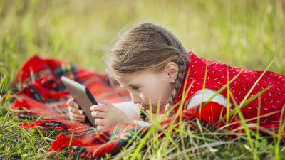 En flicka ligger på en gräsmatta på en filt och spelar på en I-Pad.