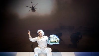 En skådespelare i vitt (Hellen Willberg) leker med ett par solglasögon medan en stridshelikopter och soldater projiceras på väggen bakom henne. 