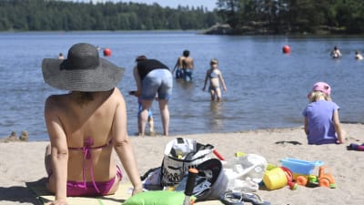 En kvinna sitter på en badstrand i bikini och hatt, i bakgrunden simmar barn och vuxna.