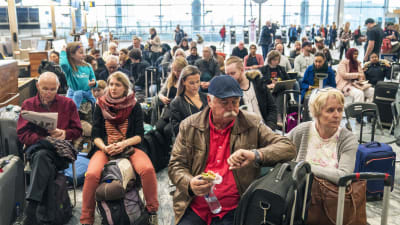 Resenärer sitter och väntar på en flygplats och en man tittar på klockan.