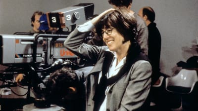 Nora Ephron står vid en filmkamera och ser glad ut.