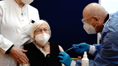Äldre kvinna med vitt hår, munskydd och glasögon får vaccin av man i blå skyddsdräkt.