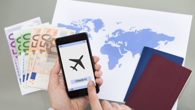 En hand håller i en mobiltelefon och ska checka in på ett flygplan. På bordet framför ligger pengar, en världskarta och två pass. 