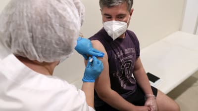 En man som bär munskydd sitter på en bänk medan en sjuksköterska vaccinerar honom.