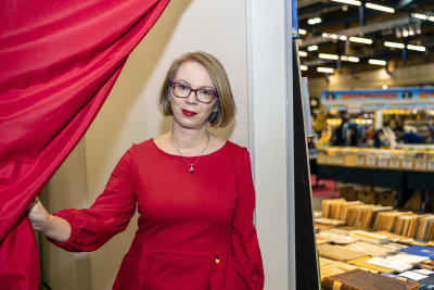 Maria Turtschaninoff tittar fram bakom en röd gardin i ett rum intill bokmässans utställningsutrymmen. 
