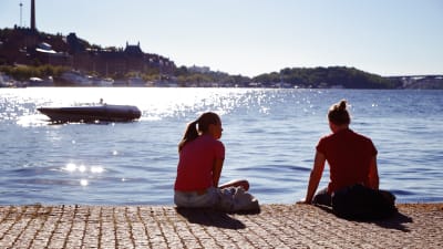 Unga människor sitter vid en kaj, i bakgrunden en båt.