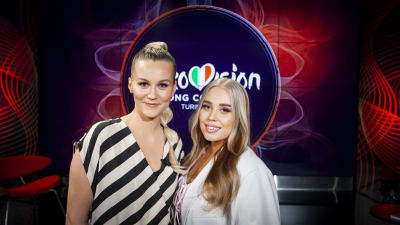 Två blonda kvinnor som ser in i kameran, ena klädd i svart-vitt randigt och den andra i vitt. I bakgrunden står det Eurovision Song Contest.