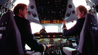 Två piloter sitter i ett flygplan vid styrpanelen. De tittar på varandra medan de sitter på varsin stol.