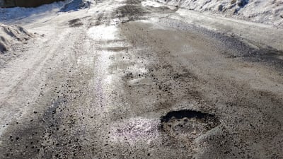 En stor grop mitt i en asfaltväg som har rejält med sand och smält snö på sig.