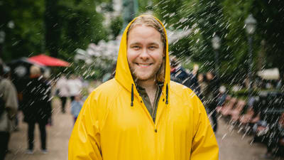 Joel Forsbacka tittar rakt in i kameran och ler brett iklädd ett gul regnjacka. Det regnar vatten runt honom.