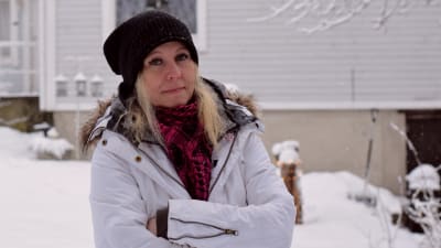 En kvinna med blont hår står utomhus i mössa och vinterkläder. Det är snö på marken.
