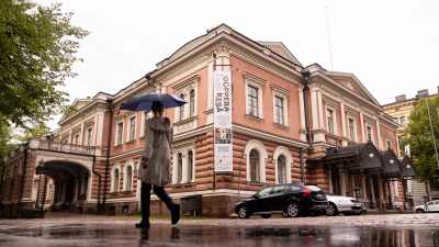 En människa promenerar på trottoaren utanför Alexandersteatern i Helsingfors. Hen har ett blått paraply.