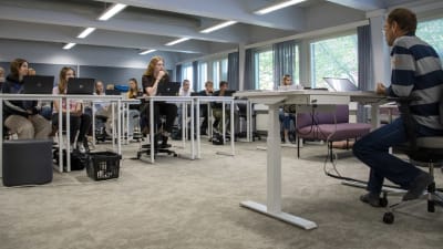 Studerande sitter bakom bord i ett klassrum. Framför dem sitter en lärare vid ett höj- och sänkbart bord.