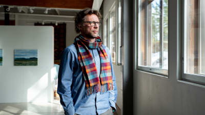Antti står i ett soligt konstgalleri och ser ut genom fönstret. Han har en blå skjorta och röd-blå-rutig halsduk på sig.
