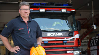 Sture Holmström med en orange hjälm i handen framför en brandbil.