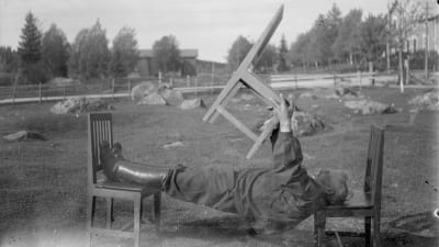 En man leker "ligga lik", där man ska hålla sin kropp så styv att man kan ligga raklång mellan två stolar och lyfta en tredje stol. Bilden är tagen 1931 i Närpes, Kalaks.