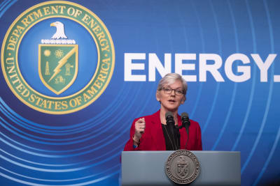 En kvinna med kort ljust hår klädd i röd kavaj står vid ett talarpodium. Bakom henne finns USA:s energiministeriums emblem och texten Energy.