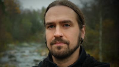Mathias Lillmåns ser in i kameran, i bakgrunden skog och en fors. Svart jacka. Långt hår i svans. Piercing i näsan.