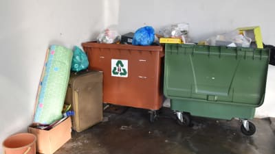 Två avfallsbehållare fyllda med skräp. Framför behållarna står en gammal kappsäck, en madrass och en låda med diverse skräp.