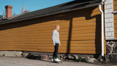 Mies skeittilaudalla, taustalla vaaleanruskea vaakalaudoitettu puutalon seinä.