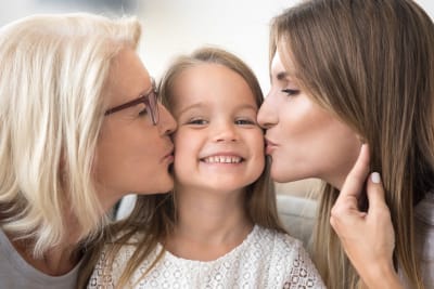 Liten leende flicka får pussar på kinden av sin mamma och mormor
