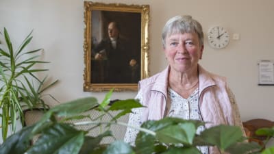 En kvinna står vid en grönväxt och tittar in i kameran.