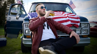 Hannes njuter av en öl framför sin Chevrolet med amerikanska flaggor i dörrarna och på motorhuven.