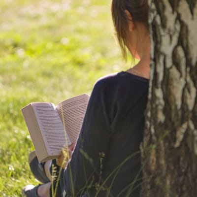 Nainen lukee kesällä ulkona kirjaa koivuun nojaten.