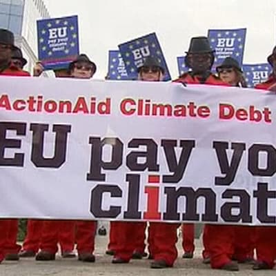 Punaisiin pukuihin ja aurinkolaseihin pukeutuneita mielenosoittajia seisoo rivissä Brysselissä. Pitävät käsissään lakanaa, jossa lukee "EU pay your climate debt".