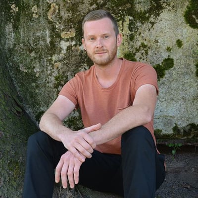 Kuvassa Henrik Blomqvist istuu kahden puunrungon välissä, taustalla vanhaa muuria