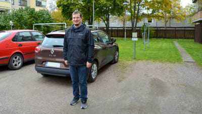En man står på en parkeringsplats bakom en brun bil.
