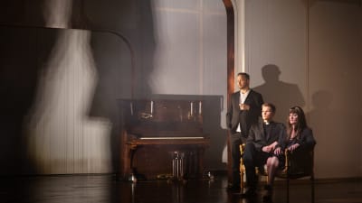 Tre människor i svart på en dunkel scen med vita väggar bredvid ett antikt piano i mörkt trä.