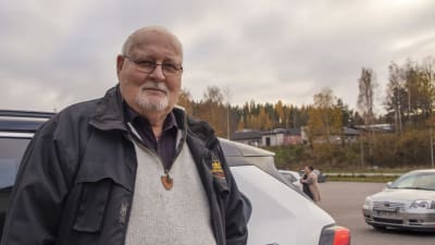 En man står vid sin bil på ett parkeringsområde och tittar in i kameran.